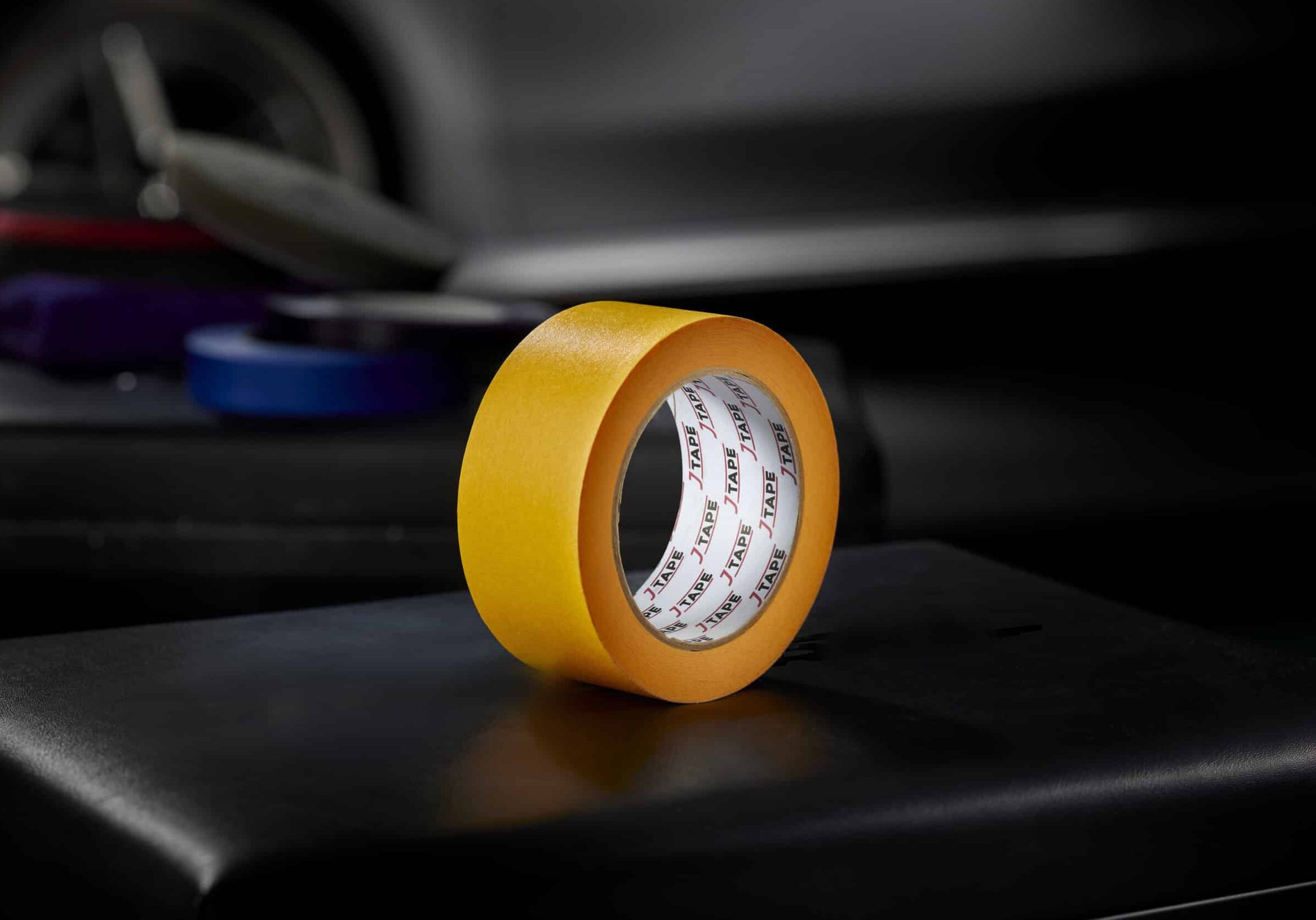A roll of bright orange wide tape, standing upright on a dark surface, with a blurred background that includes another roll of tape and a car door.